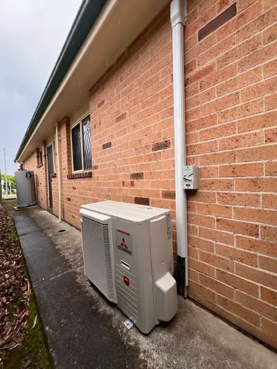 Outdoor condenser beside a brick wall with conduit running straight up to the eaves.